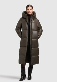 Palton pufos lung de culoare verde măsliniu, cu glugă, prezentând un design matlasat, fermoar frontal și buzunare laterale. Purta peste un pulover negru cu guler înalt.