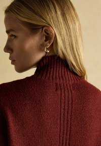 Burgunderfarbener Strickpullover mit hohem, geripptem Kragen und strukturiertem Stoff. Goldene Ohrringe sind sichtbar und setzen einen Kontrast zum Outfit.