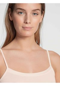 Top a canotta rosa chiaro con spalline sottili, realizzato in un tessuto liscio e elasticizzato. La modella ha i capelli castani lisci e un trucco minimale.