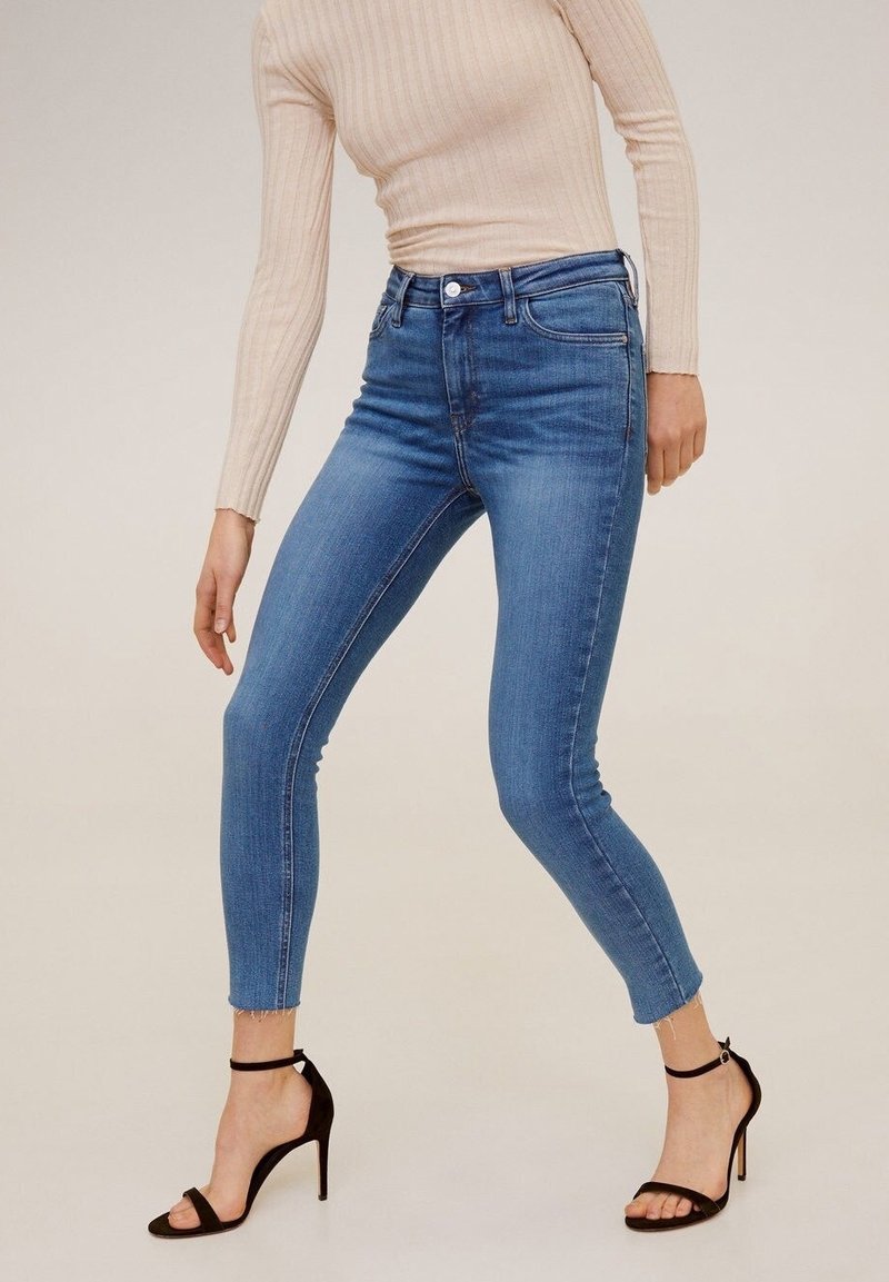 Personne portant un jean skinny bleu, un haut beige à manches longues côtelé et des sandales noires à talons hauts sur un fond uni.