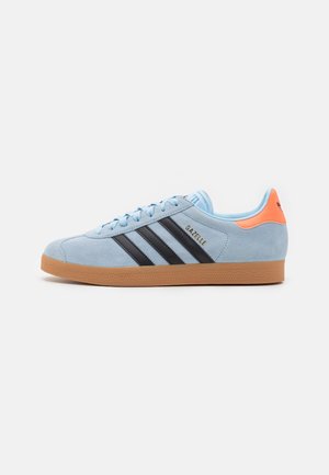 Baskets Adidas Gazelle bleu clair avec bandes noires, semelle en gomme et languette orange au talon sur fond blanc, vue de côté.