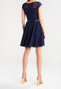 Robe bleu marine avec un corsage en dentelle et des manches courtes, jupe évasée, ceinture en satin et texture lisse. Portée avec des sandales à talons.