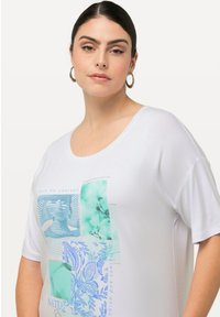 Camiseta blanca de manga corta con un estampado gráfico multicolor que incluye texto y patrones florales. Hecha de material suave, tiene un corte holgado.
