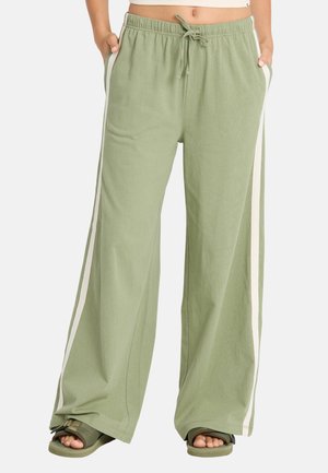 Persoon draagt losse lichtgroene broek met witte zijstrepen, elastische tailleband, trekkoord, handen in de zakken en groene sandalen met open tenen.
