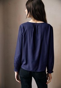 Blusa blu navy a maniche lunghe con scollo arricciato e polsini delle maniche plissettati, realizzata in un tessuto liscio.