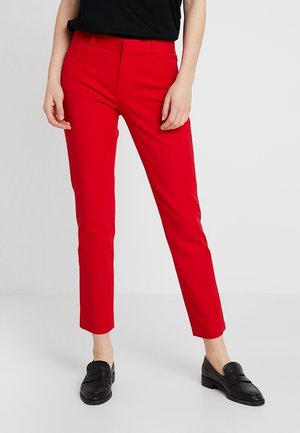 Mujer con pantalones tobilleros ajustados de color rojo brillante, top negro y mocasines negros sin cordones, de pie frente a un fondo claro y liso.