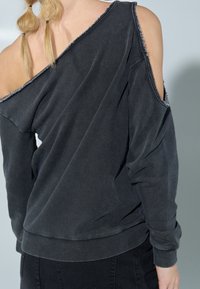 Sweat-shirt noir à épaules dénudées en tissu doux avec bords effilochés et coupe ample. Poignets et ourlet côtelés ; porté avec un pantalon foncé.