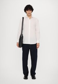 Camisa de botones blanca de manga larga, jeans azul oscuro, zapatos de cuero negros y un bolso negro de hombro, de pie contra un fondo sencillo.