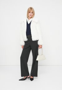 Giacca in pelliccia sintetica bianca, maglione blu navy a scollo a V, pantaloni grigi a righe a gamba larga, décolleté neri con cinturino, piccola borsa beige. Sfondo bianco.