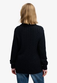 Maglione blu navy lavorato a maglia con texture a coste e motivo a trecce. Presenta un collo alto, spalle scese ed è abbinato a una maglietta rosa sotto.