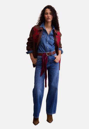Donna in piedi che indossa una giacca marrone ricamata sopra una camicia di jeans e jeans, con cintura marrone e sciarpa bordeaux legata in vita.