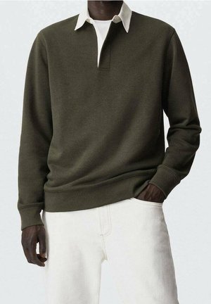 Homme portant un pull vert foncé à manches longues avec une chemise col blanc en dessous et un pantalon de couleur claire, main dans la poche.