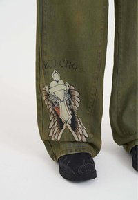 Grüne Jeanshose mit einem großen Grafikdesign eines Vogels mit Flügeln und einem dekorativen Element an einem Bein. Auffällige Nähte und eine lockere Passform.