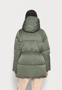 Grön puff-jacka med huva, som har quiltade sektioner, cinchade midja och slät textur, visad från baksidan på en modell.