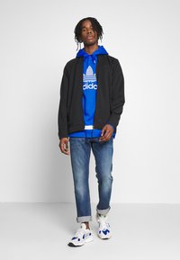 Černá zipová bunda přes modrou mikinu s bílým logem adidas, v kombinaci s světle modrými džíny a bílými teniskami s modrými akcenty.
