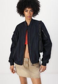 Zwarte bomberjack met een ritssluiting aan de voorkant, geribbelde manchetten en een zak op de linkermouw. Draagt over een cropped rood top en een beige rok.