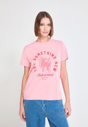 Camiseta de algodón rosa con mangas cortas que presenta un gráfico de leopardo y detalles florales, resaltados por texto en rojo. Combinada con pantalones vaqueros azules.