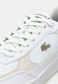 Λευκά δερμάτινα αθλητικά παπούτσια με ανοιχτό γκρι σουέτ λεπτομέρειες, επίπεδα κορδόνια και χρυσό λογότυπο Lacoste. Διαθέτουν περφορένσεις στο πλάι για καλύτερη αναπνοή.