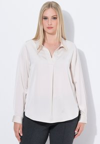 Blusa a maniche lunghe color beige chiaro in tessuto lucido con scollatura a V, pieghe sottili e orlo curvo. Presenta un colletto classico e maniche con polsini.