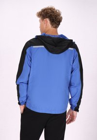 Giacca leggera blu con dettagli neri, cappuccio regolabile, polsini elastici e striscia riflettente sulle spalle. Materiale liscio e resistente all'acqua.