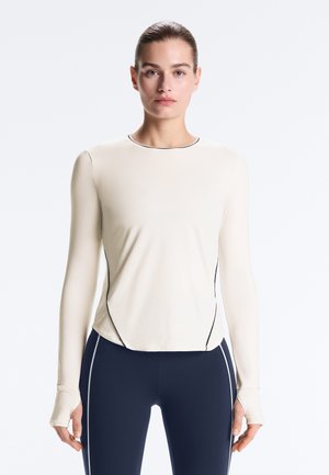 Frau mit zurückgebundenem Haar, die ein enganliegendes langärmliges cremefarbenes Oberteil mit schwarzer Paspelierung und marineblaue Leggings mit weißen vertikalen Streifen trägt.