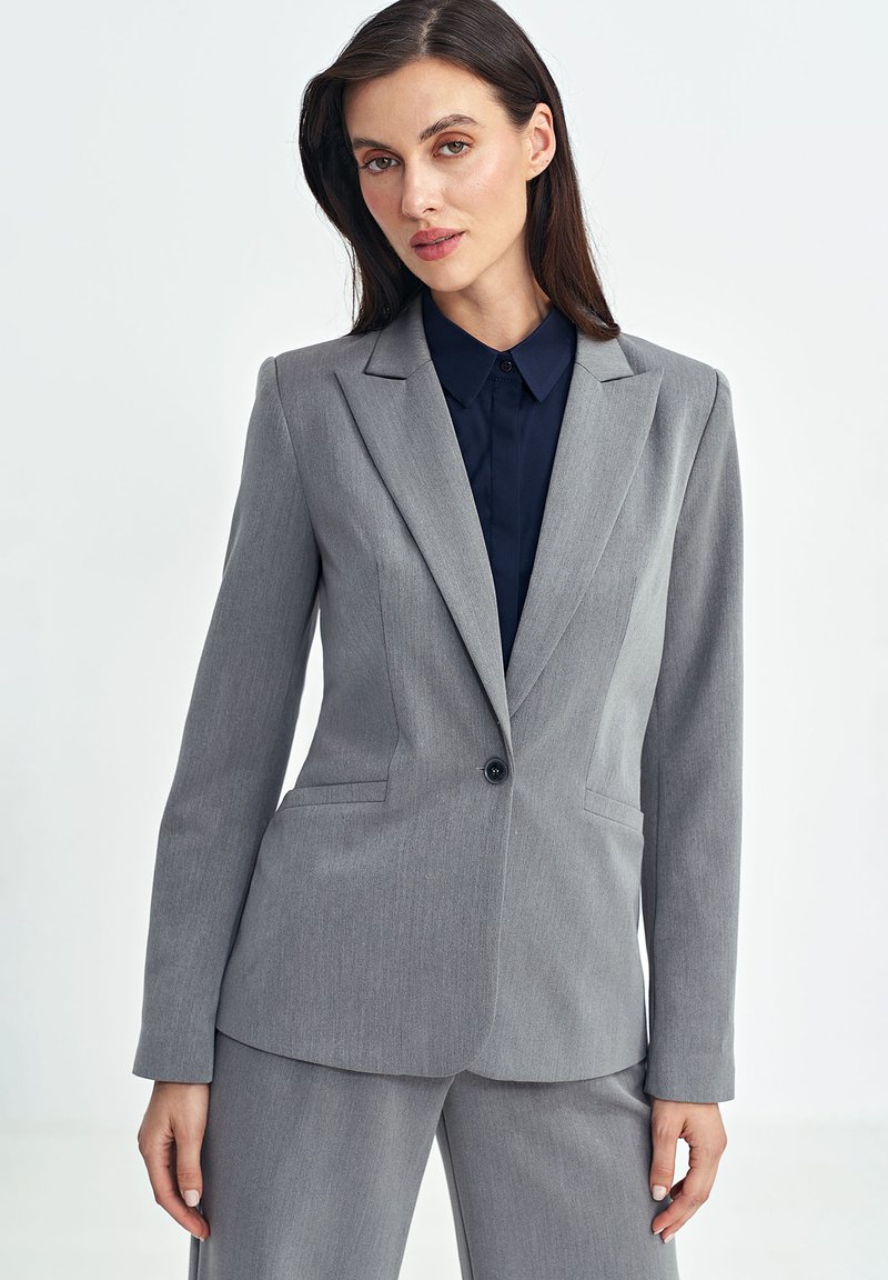 Szary, dopasowany żakiet z klapami i pojedynczym zapięciem na guzik. Noszony na ciemnoniebieskiej koszuli, z dwiema bocznymi kieszeniami. Gładka tekstura.
