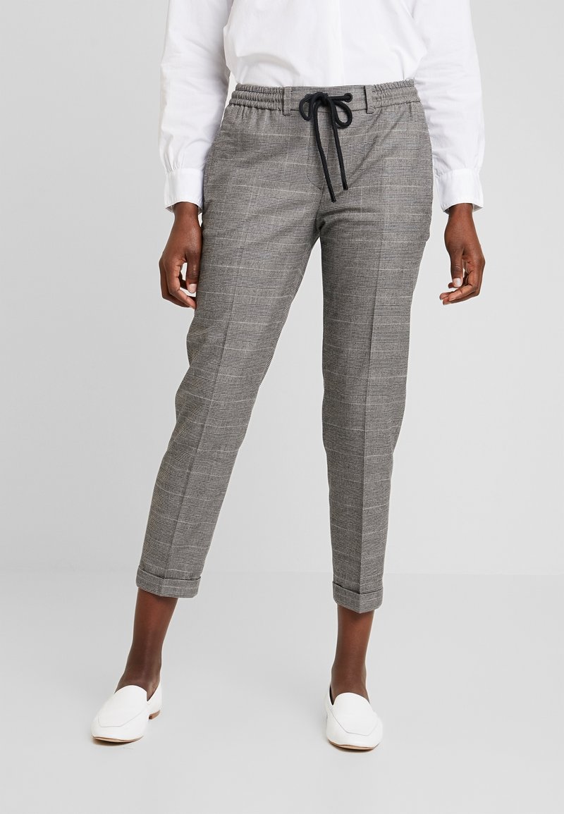 Personne portant un pantalon court gris à carreaux avec cordon noir et des mocassins blancs, debout sur un fond gris clair.