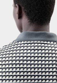 Polo a pattern geometrico bianco e nero con colletto a coste grigio, caratterizzato da piccoli triangoli in colori alternati e tessuto testurizzato.