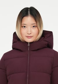 Maroon puffer jacket with a high collar, full zipper, and quilted texture. Features a lined hood and subtle sheen to the fabric.