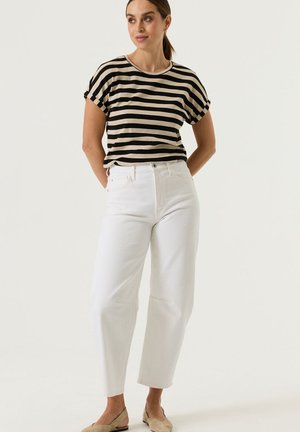 Vrouw staat met handen achter rug, draagt een zwart-beige gestreepte top met korte mouwen, witte broek met hoge taille en beige platte schoenen.