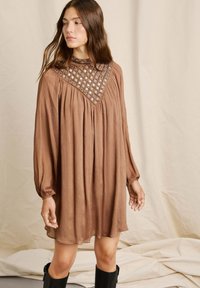 Vestido marrón hecho de un tejido liviano, con mangas largas, un escote recogido y un intrincado bordado alrededor del cuello.