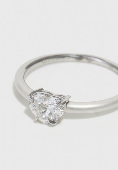 Anillo de plata con una banda lisa y un engaste de piedra en forma de doble corazón, que muestra superficies facetadas y claras, así como acentos metálicos.