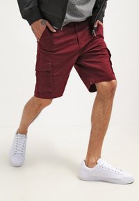 Shorts cargo bordeaux avec poches latérales, fabriqués en tissu léger. Associés à des baskets blanches avec une semelle texturée et un design à lacets.