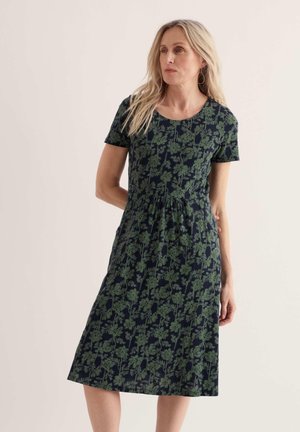 Frau mit langen blonden Haaren trägt ein marineblaues knielanges Kleid mit grünem Blumenmuster, kurzen Ärmeln und geraffter Taille, stehend vor einfarbigem Hintergrund.
