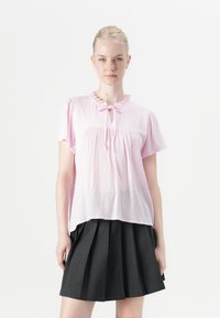 Blusa rosa chiaro a maniche corte con scollo arricciato, dettaglio a occhio di bue e tessuto texturizzato abbinata a una gonna nera a pieghe.