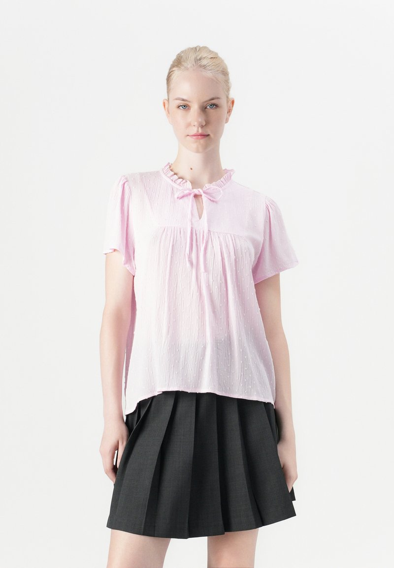 Blusa rosa chiaro a maniche corte con scollo arricciato, dettaglio a occhio di bue e tessuto texturizzato abbinata a una gonna nera a pieghe.