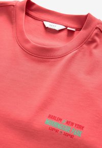 Koralfarvet crew-neck t-shirt lavet af bomuld med en glat tekstur. Har trykt tekst i turkis og rød: "HARLEM / NEW YORK MORNINGSIDE PARK."
