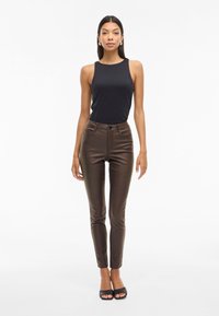 Hnědé skinny kalhoty z umělé kůže spárované s černým upnutým topem bez rukávů. Hladká textura a design s pěti kapsami; modelka má na sobě černé sandály na podpatku s otevřenou špičkou.