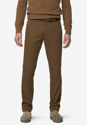 Mann mit braunen enganliegenden Hosen, einem passenden braunen Pullover, einem bunten gewebten Gürtel und hellbraunen Wildlederschuhen steht vor einem schlichten Hintergrund.