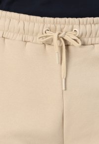 Los pantalones de chándal beige presentan una cintura elástica con un cordón funcional. La tela tiene una textura suave con pequeños detalles de costura.