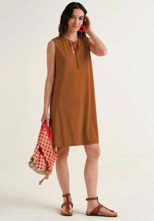 Donna con vestito marrone senza maniche e orecchini rossi vistosi, che tiene una borsa decorata, in piedi con sandali su sfondo uniforme.