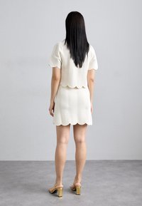 Ensemble composé d'un haut crème à bord festonné et d'une jupe, présentant des motifs ondulés texturés. Assorti à des sandales à talons dorés. Fond gris neutre.