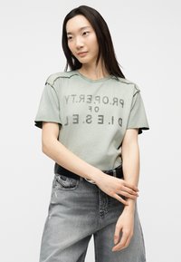 Νεαρή γυναίκα με μακριά σκούρα μαλλιά φοράει μια ξεθωριασμένη πράσινη φαρδιά μπλούζα και γκρι ξεβαμμένα τζιν, στεκόμενη με τα χέρια σταυρωμένα πάνω στο χέρι.