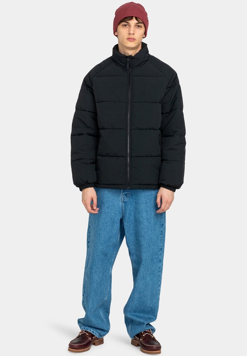 Czarna kurtka puchowa z wysoką kołnierzem, poziomym przeszyciem i zapięciem na zamek, zestawiona z luźnymi niebieskimi dżinsami i brązowymi butami.