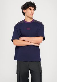 Navy blue T-shirt made of soft fabric, featuring a purple collar and shoulder accents, and a rectangular label with "70" on the front.