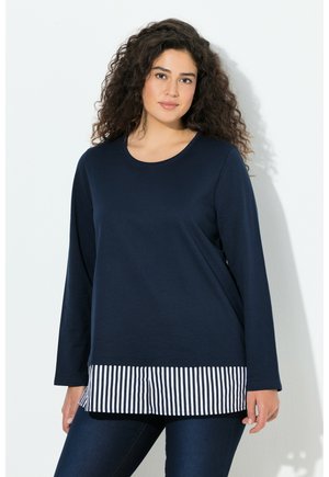 Kobieta z kręconymi ciemnymi włosami, ubrana w granatowy długi rękaw nałożony na czarną i białą pasiastą koszulę oraz ciemne dżinsy, stojąca na tle białej ściany.