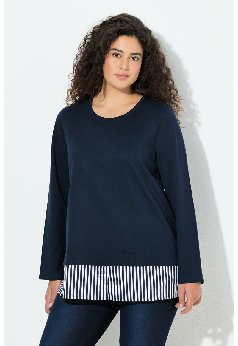 Frau mit lockigem dunklem Haar, die ein marineblaues Langarmshirt trägt, darüber ein schwarz-weiß gestreiftes Hemd und eine dunkle Jeans, stehend vor einer weißen Wand.