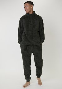 Jonge man die blootsvoets staat in een donkergroene, fuzzy fleece zip-up pullover en bijpassende jogger broek, kijkt naar links met zijn handen in zijn zakken.