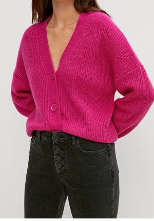 Frau trägt eine leuchtend pinke, geknöpfte Strickjacke mit tiefem V-Ausschnitt und hoch taillierte schwarze Jeans mit mehreren Knöpfen.