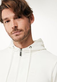Vit zip-up hoodie med en mjuk yta, justerbara dragsko och minimalistiska logotypdetaljer. Har en tajt och avslappnad design.